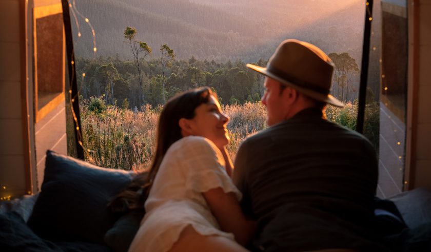 Verliebtes Paar liegt im Camper und im Hintergrund ist durch die geöffnete Hecktür eine weite bewaldete Landschaft im Sonnenuntergang zu sehen