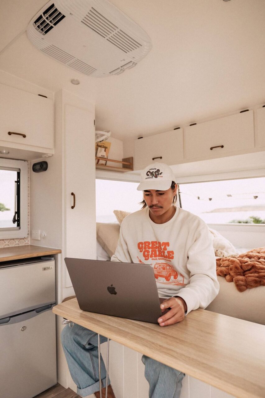 Ein Mann mit Capy sitzt im Wohmobil und schaut aus sein MacBook